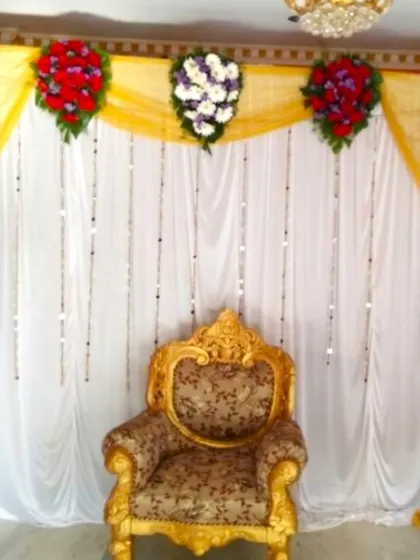 A simple flower decoration for a Half Saree ceremony. The backdrop of yellow and white drapes is accented with colorful flower bouquets and a traditional gold-colored chair.