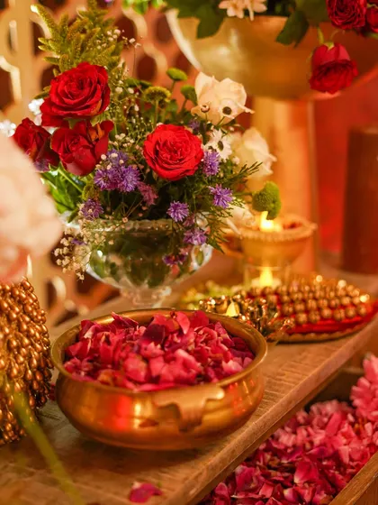 A close-up of a brass bowl filled with fresh rose petals, surrounded by smaller floral arrangements and the warm glow of a diya.