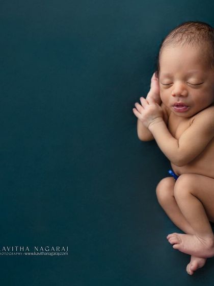 A simple, minimalist portrait of a newborn against a deep teal backdrop. The focus is entirely on the baby's natural, curled-up pose and peaceful expression.