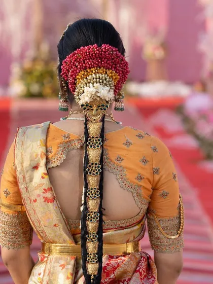 A stunning view of a bride's back, showcasing the scalloped cutout of her orange blouse and elaborate hair adornments. The blouse embroidery perfectly matches the traditional aesthetic of her wedding look.