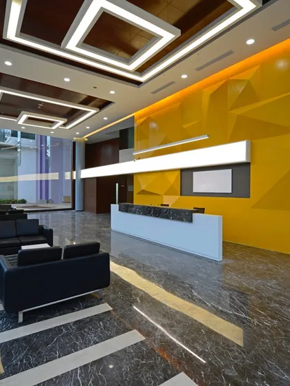 A grand, double-height lobby with a bold, geometric yellow feature wall. The polished marble floors and sophisticated ceiling design with recessed lighting create a luxurious and impressive entrance for a corporate building.