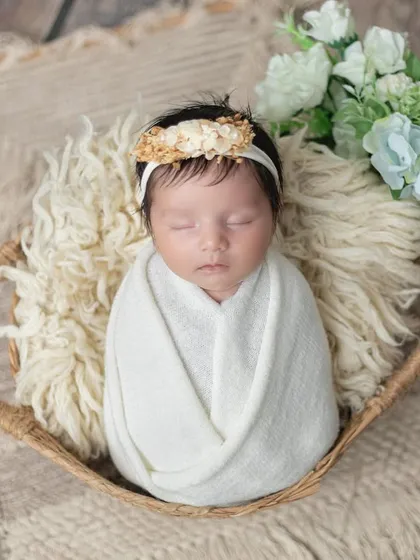 A newborn girl wrapped in white, resting in a rustic basket with soft flowers.