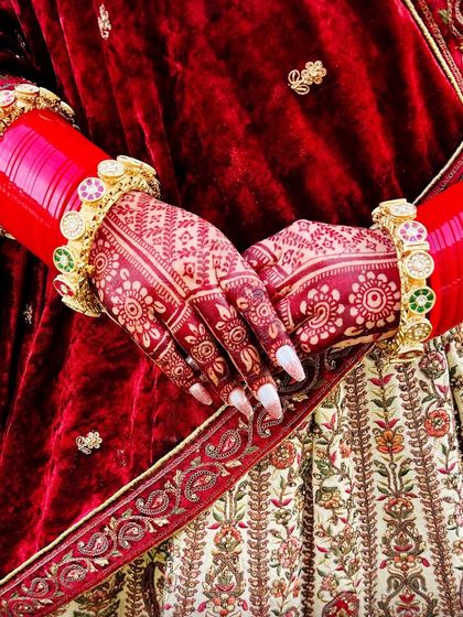 A close up of a bride's hands, showing the beautiful floral mehndi stain against her red and cream lehenga. The color is a perfect, classic bridal red.