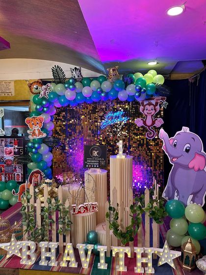 A jungle-themed party with a touch of sparkle. This setup combines a gold shimmer wall with a green and blue balloon arch, cute animal cutouts, and illuminated letters for the name.