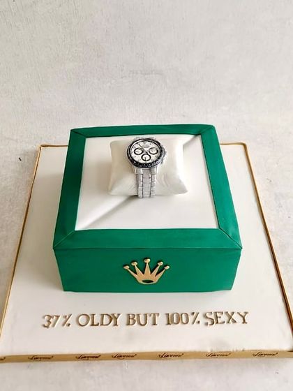 A luxurious Rolex-themed cake. The green watch box is the cake, and it's topped with a detailed, edible Rolex watch resting on a sugar pillow. A perfect cake for a milestone birthday.