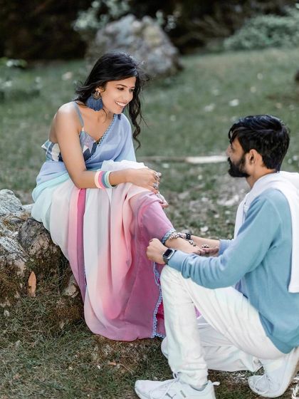 A sweet, romantic gesture in a park. The groom kneels to adjust the bride's anklet, a modern-day Prince Charming moment captured in a beautiful, natural setting.