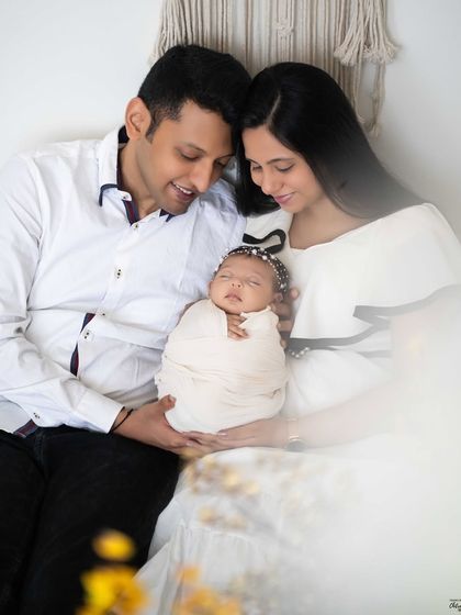 A cozy and relaxed family portrait. Seated together, the parents create a loving frame around their sleeping newborn, making for a beautifully composed memory.