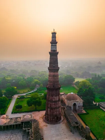 Qutub Minar stands tall against a hazy sunrise in this aerial shot. The soft morning light accentuates the texture of the historic minaret and the surrounding green complex, offering a serene view of this Delhi landmark.