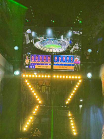 Another view of the IPL screening setup. The LED screen clearly displays the match scores and visuals, while the bright yellow lights on the truss create a stadium-like feel.