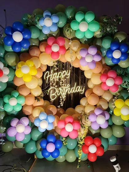 A unique birthday backdrop made of a full circle of balloons with colorful balloon flowers. The "Happy Birthday" neon sign in the center makes it a cheerful and eye-catching decoration.