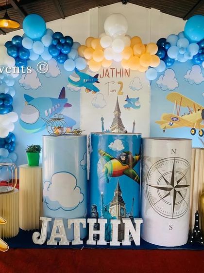 An adventurous airplane theme for a 2nd birthday. The decor includes a sky-print backdrop, plane cutouts, and pedestals with compass and cloud designs.