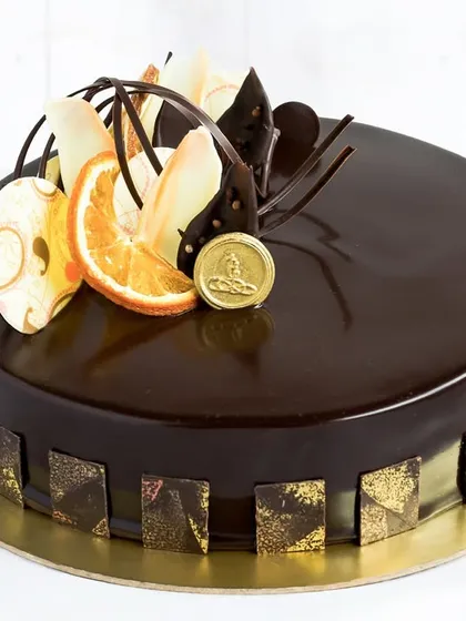 Our Chocolate Orange Cake, a sophisticated creation with a deep chocolate mirror glaze. It's decorated with candied orange slices and abstract white and dark chocolate pieces, offering a classic flavour pairing with an artistic touch.