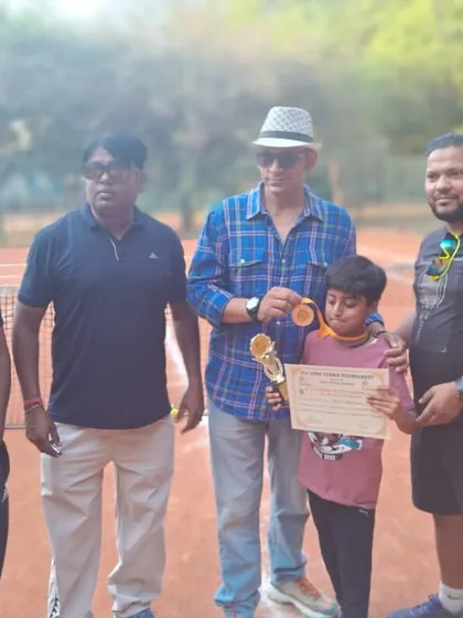A young player receiving his medal and certificate for the U12 Aptitude Title at the Ideal Tennis Open Tournament, surrounded by coaches and officials on the clay court.