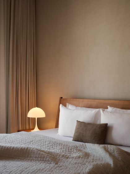 A close-up of the bedside in a minimalist bedroom, featuring a simple wooden headboard, crisp white linens, and a warm, glowing lamp.
