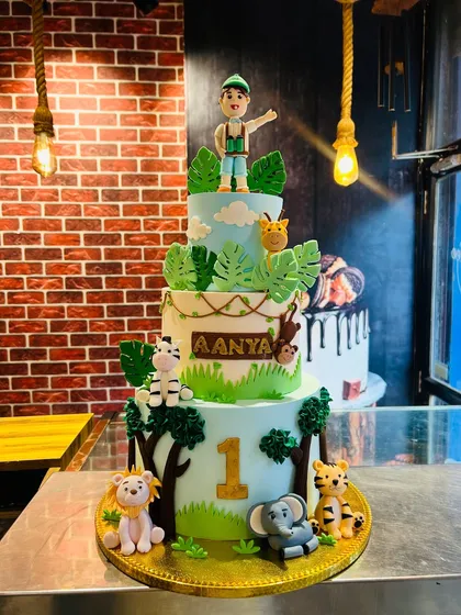 A grand three-tier jungle safari cake designed for a first birthday party. It features a variety of handcrafted fondant animals like a lion, elephant, zebra, and a little explorer figurine, all made from 100% vegetarian ingredients.