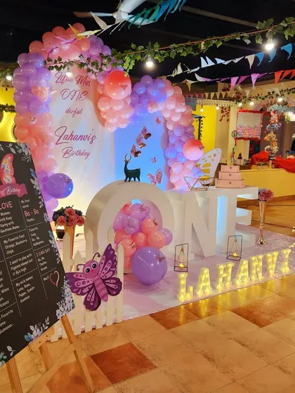 A "Love is ONEderful" first birthday party. This setup includes a large "ONE" marquee, a custom milestone chalkboard, and a beautiful butterfly and balloon backdrop in pink and purple.