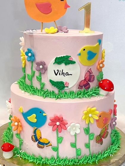 A cheerful two-tier cake with a birds and garden theme for a first birthday. The cake is decorated with colorful birds, flowers, and butterflies.