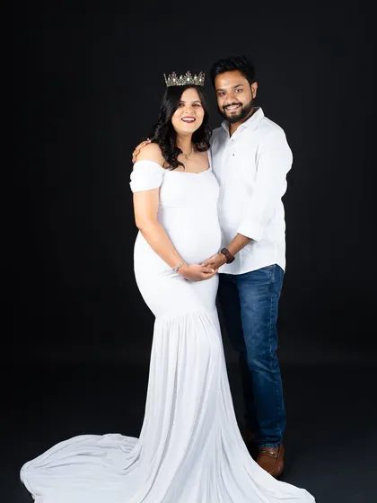 A classic couple's portrait against a solid black background. The mom-to-be in a white gown and crown stands with her partner, creating a timeless and elegant image.