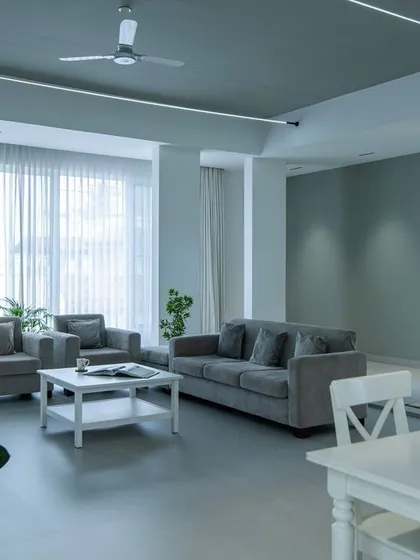 A serene living room where shades of grey create a calm, unified atmosphere. The open layout connects the seating area to the dining space, unified by clean flooring and linear ceiling lights.