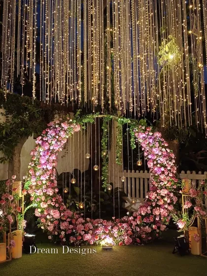 The circular floral backdrop illuminated at night. The combination of the pink flowers, hanging glass globes, and the warm fairy lights creates an enchanting photo opportunity.