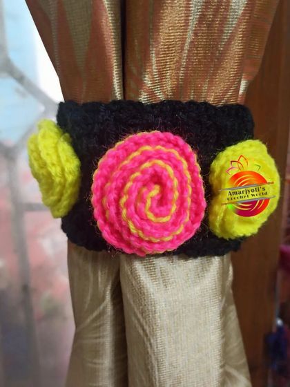 A modern and vibrant curtain tie-back design. It features a black band decorated with a central pink and yellow swirl and two bright yellow pom-poms on the sides.