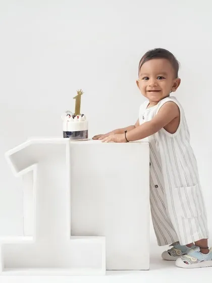 A little boy proudly stands next to a large '1' prop and his first birthday cake in a clean, minimalist all-white studio setting.