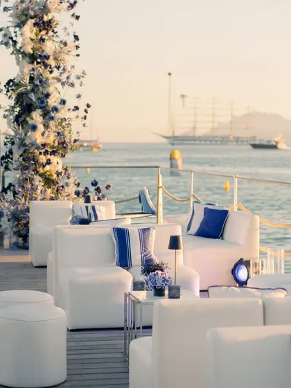 The lounge setup for the welcome dinner, with comfortable white sofas, blue and white striped cushions, and a floral arch, all set against the golden hour glow on the water.