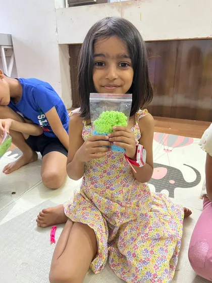 A young girl proudly holds up the colorful, textured material she created in a workshop. The pride in her creation is evident.