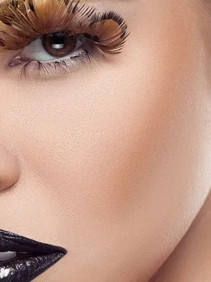 A macro shot of the model's skin and eye, showing the intricate detail of the feather makeup. The lighting reveals natural skin texture while maintaining a polished, high-end feel.