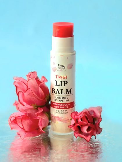 This tinted lip balm is surrounded by fresh rose petals against a reflective, water-like surface. The setup beautifully visualizes the "rose oil" ingredient and creates a refreshing, hydrating feel.