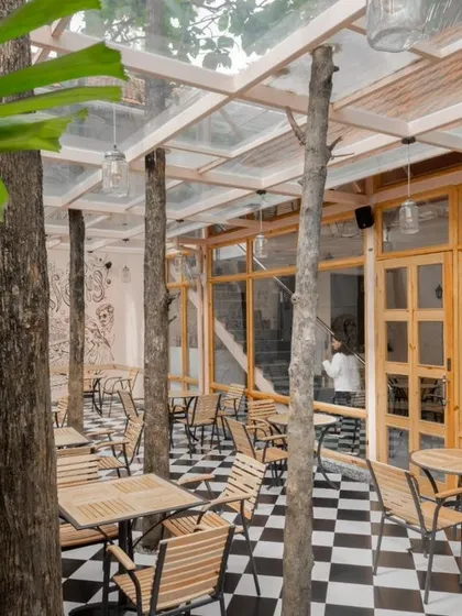 The outdoor seating area is designed to fit between the trunks of mature trees. The checkered floor and simple furniture create a classic European cafe feel, transported into a tropical Bangalore garden.