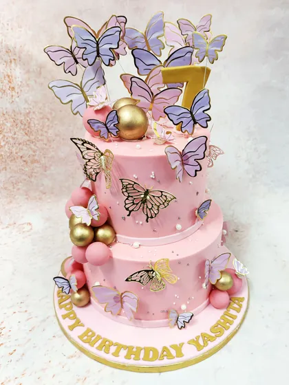 This magnificent two-tiered pink butterfly cake is a flight of fancy, perfect for a truly special birthday. A whirlwind of delicate butterflies in shades of lilac and gold flutter around the soft pink tiers, which are adorned with shimmering gold and pink spheres, creating a sense of movement and enchantment.