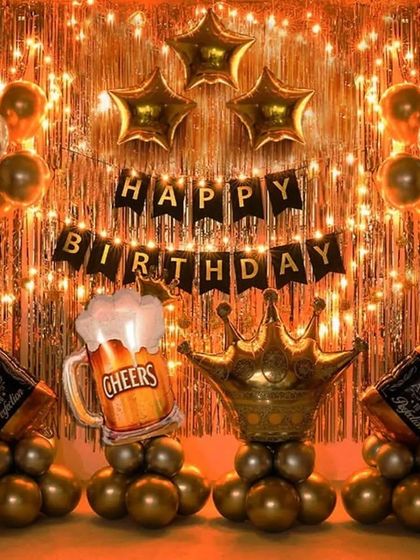 A birthday party setup with a "Cheers" theme, using a gold foil curtain, star balloons, and large foil balloons shaped like a beer mug, crown, and whiskey bottles.