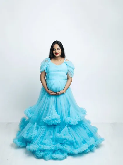 A beautiful solo maternity portrait featuring a stunning, layered blue gown. The clean white studio background makes the color pop, creating a bright, airy, and elegant photograph that celebrates the mother-to-be.