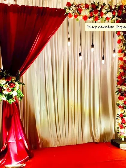 A versatile backdrop with a red and white theme, suitable for anniversaries, engagements, or even corporate events. The design features an asymmetrical floral arch and hanging Edison bulbs.