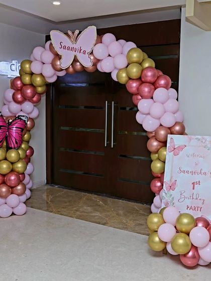An elegant entrance for a butterfly-themed first birthday, with a gold, rose gold, and pink balloon arch and a personalized welcome sign.