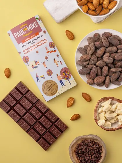 A flatlay of a dark chocolate bar with its ingredients, cocoa beans and almonds, on a cheerful yellow background. This style is modern, graphic, and full of energy.