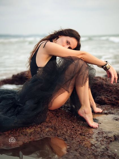 A close-up, moody portrait of a model in a black dress, seated on the rocks. The composition is intimate and emotional, with the ocean waves in the background.