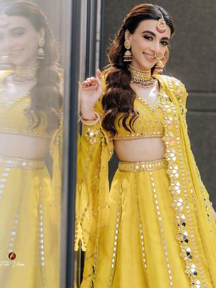 A joyful bride in a vibrant yellow mirror-work lehenga, her smile reflected in the glass beside her, creating a bright and happy portrait.