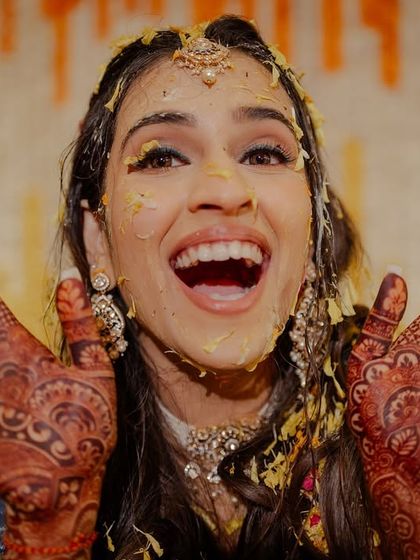 "Ta-da!" A bride joyfully shows off her mehndi-covered hands, even while covered in haldi paste. Her expression is priceless.