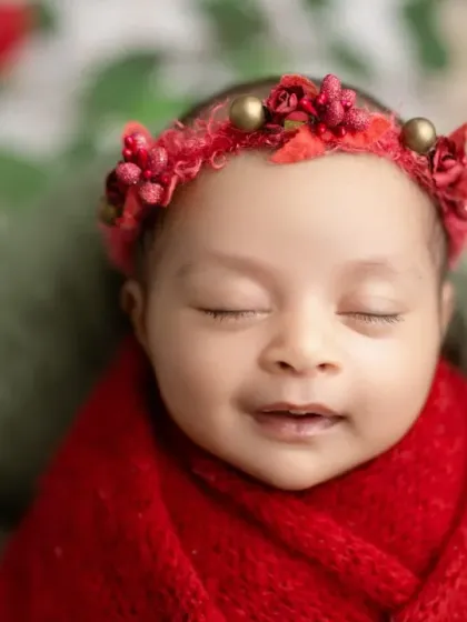 A close-up of a smiling newborn in a red wrap, with a matching red floral headband.