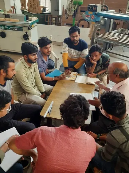 A huddle of students gathers around an instructor for a focused lesson, examining small wooden components and discussing joinery techniques.