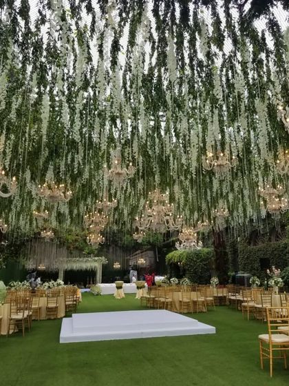 A majestic reception hall where greenery and white florals hang from the ceiling like a natural canopy. Dozens of crystal chandeliers are interspersed throughout, casting a magical glow over the entire space.