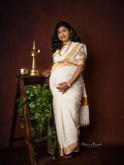 A serene portrait featuring a traditional lamp (diya) and greenery. The mom-to-be's classic white and gold saree completes this elegant, cultural look.