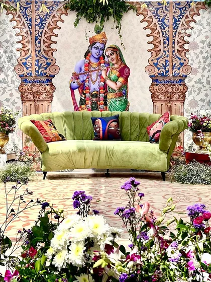 A divine Mehendi seating area with a backdrop of Radha and Krishna, surrounded by lush florals, perfect for a devotional theme.