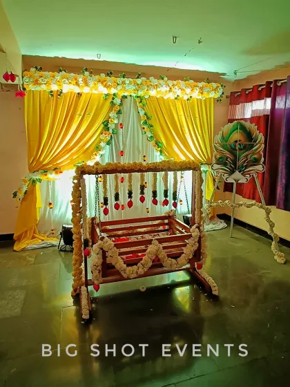 A traditional cradle ceremony with a touch of yellow and white. The backdrop features bright yellow drapes, and the wooden cradle is decorated with delicate flower strings. A peacock feather prop adds a symbolic and auspicious element to the decor.