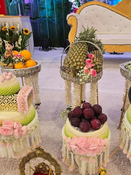 An elegant display of fresh fruits and traditional Thai floral arrangements on decorative stands for a baby shower.