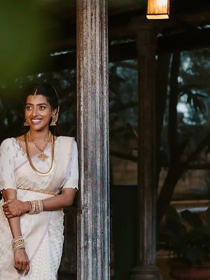 A bride in a simple white saree leans against a weathered pillar, the evening light creating a soft, romantic mood.