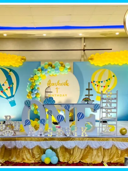 A breathtaking hot air balloon theme for a first birthday. This expansive stage setup includes a large 'ONE' prop, multiple hot air balloon models, cloud and sky backdrops, and extensive balloon work in a cheerful blue and yellow palette.