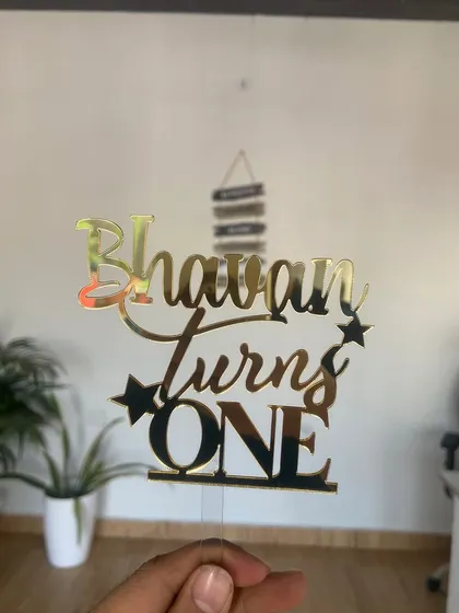 A "Bhavan turns ONE" topper in gold acrylic, featuring a mix of cursive and bold fonts with star accents for a celebratory feel.